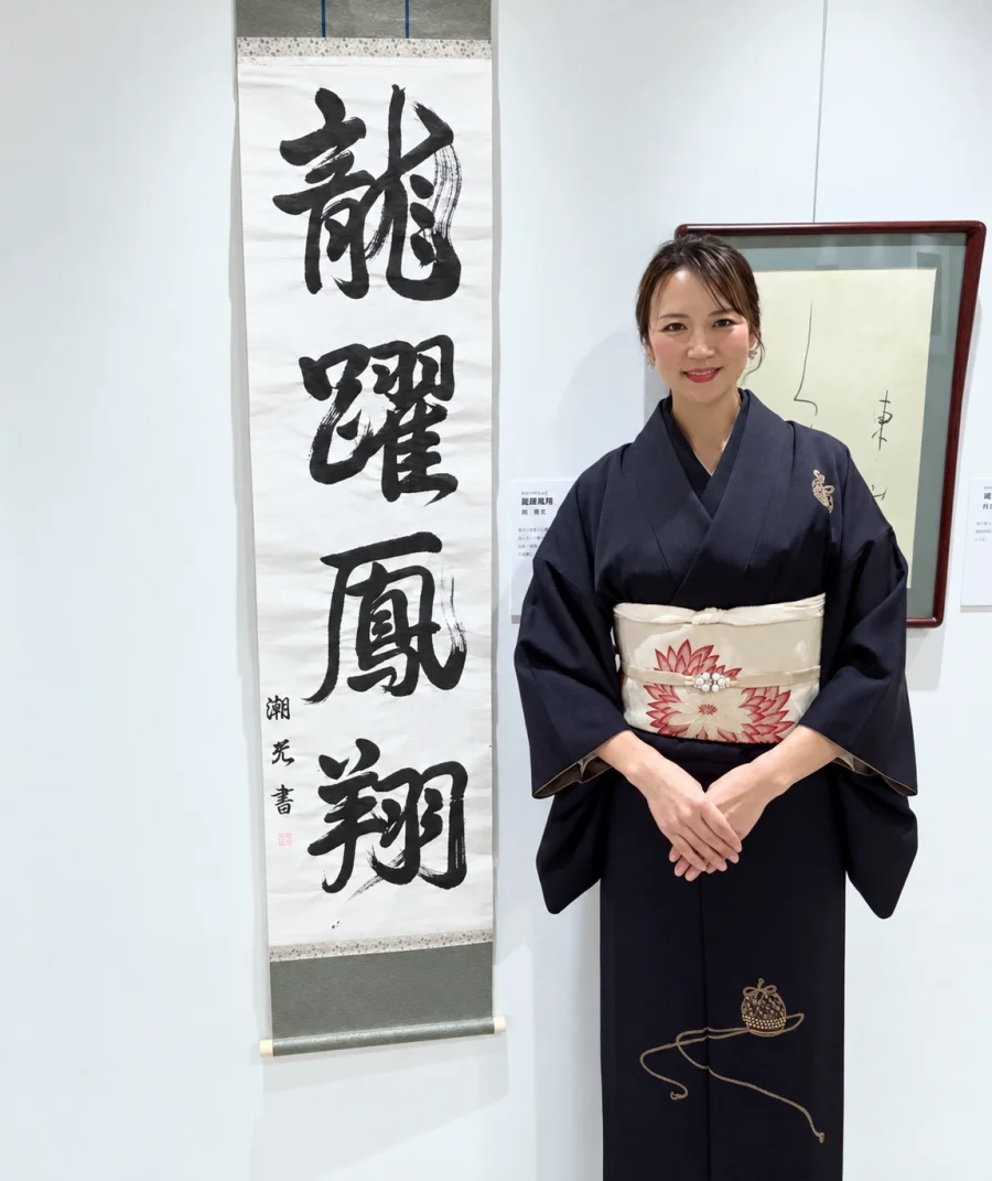 展覧会で、掛け軸の書作品「龍躍鳳翔」の横に立つ谷 弥生さん